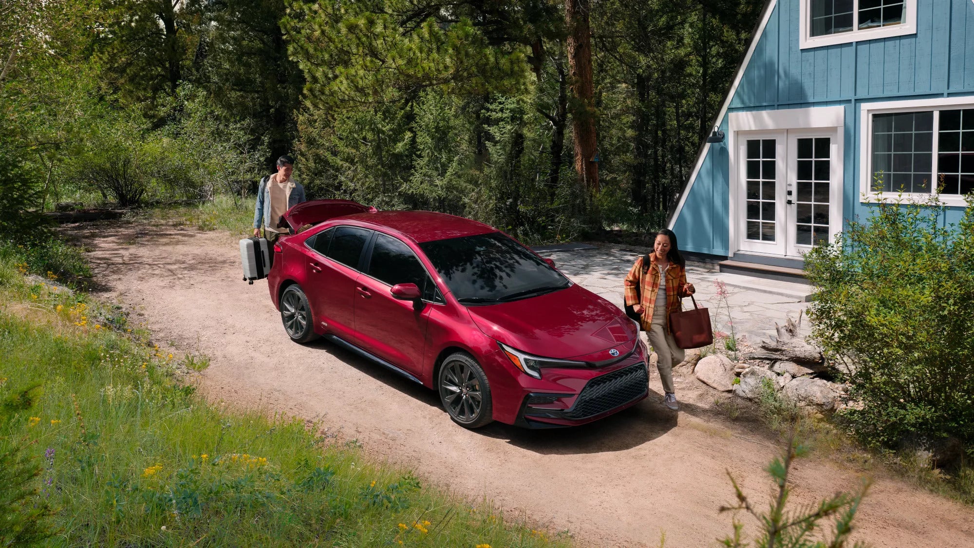a toyota corolla parked next to a cabin in the woods with 2 people unpacking