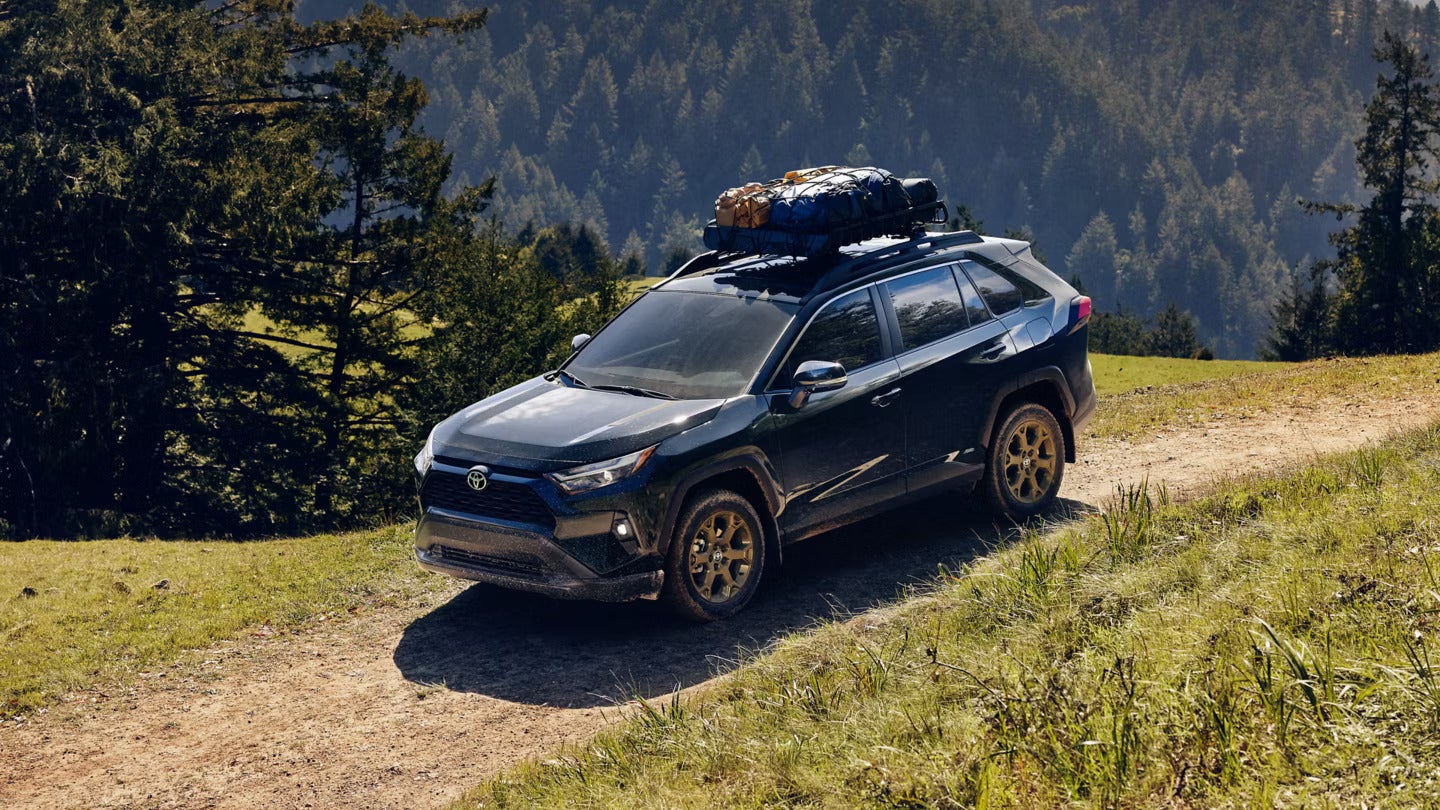 a blue 2025 toyota rav4 driving on a dirt path in teh woods with camping gear in the roof rack.