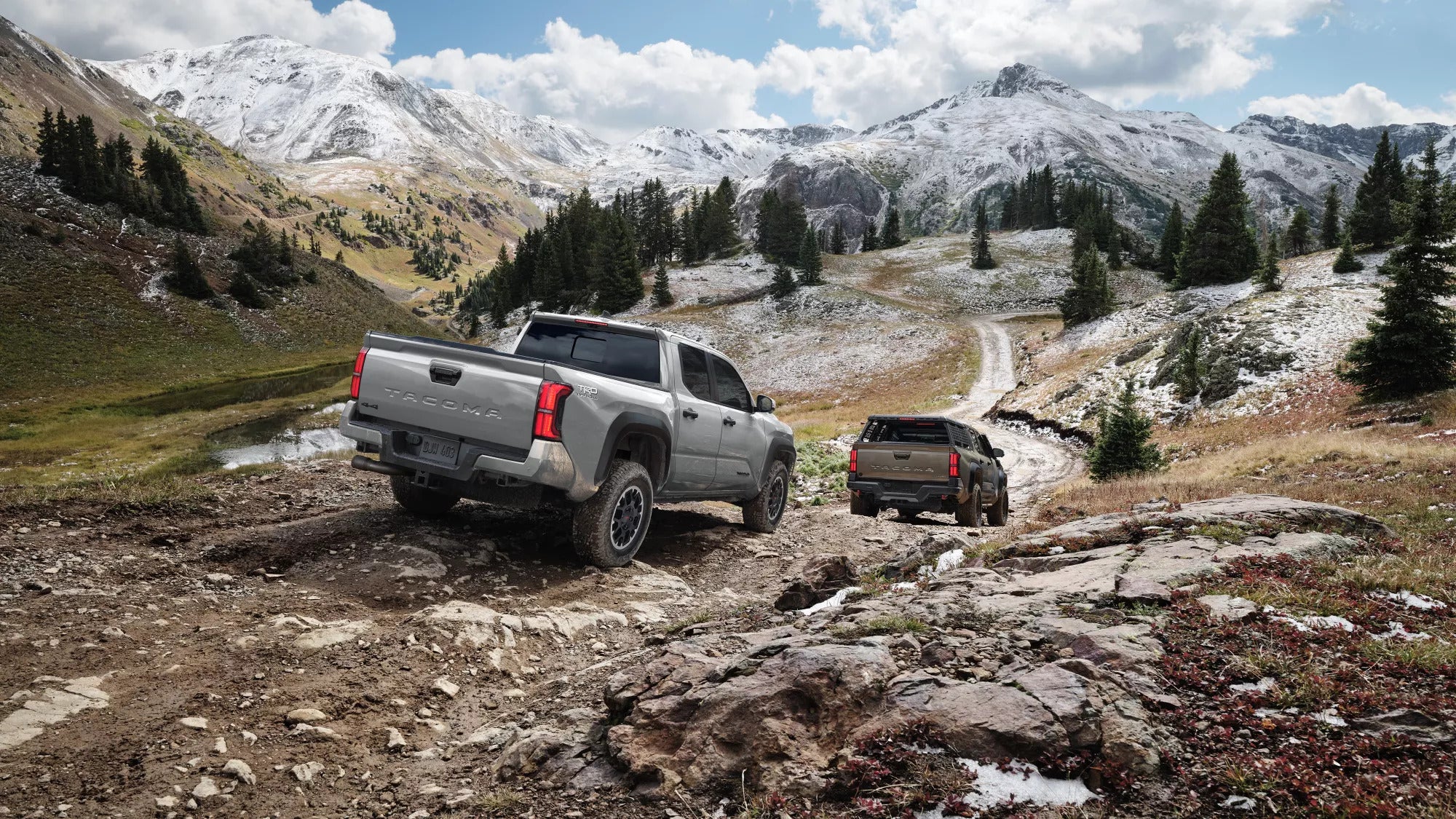 Two Toyota Tacoma trucks driving on a rugged, snow-capped mountain trail.