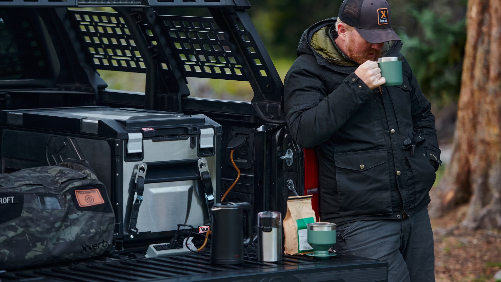 Man enjoying a warm drink by a Toyota Tacoma Trailhunter's equipped truck bed setup for outdoor adventures.