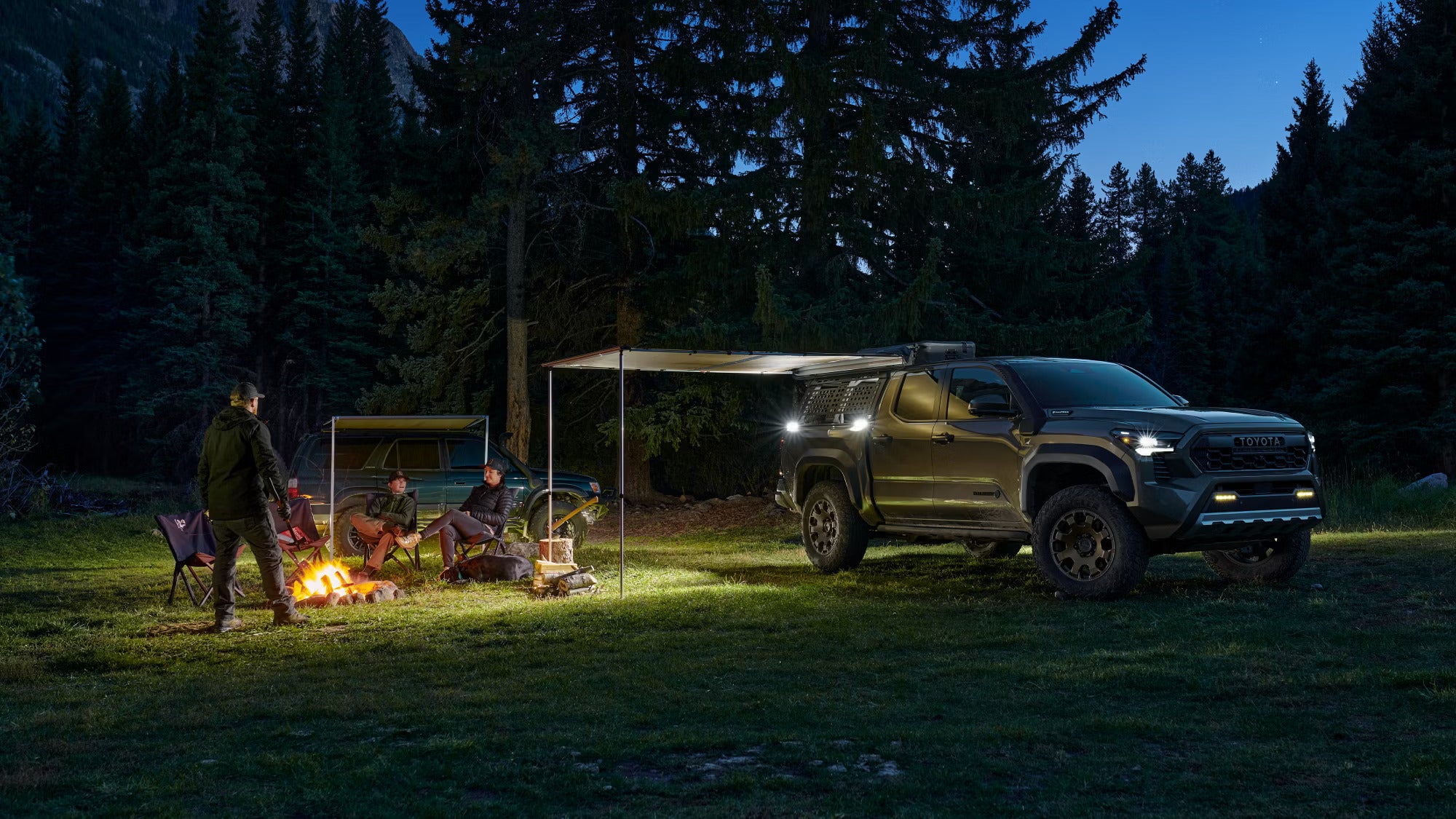 Campers relaxing by a fire with a Toyota Tacoma Trailhunter equipped with an awning at a forest campsite.