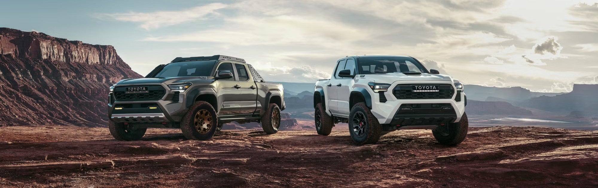 Two Toyota pickup trucks parked on rugged terrain with a scenic canyon backdrop under a partly cloudy sky.