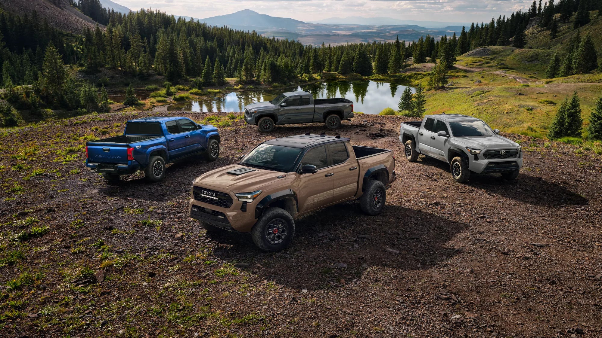 Four 2025 Toyota Tacoma trucks in different colors parked near a scenic lake surrounded by lush greenery and mountains.